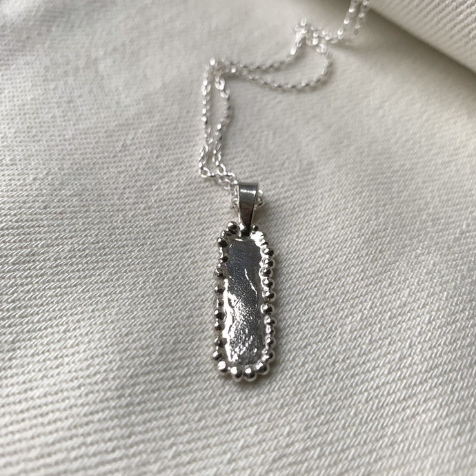 an oblong bar silver pendant necklace on a silver chain, with molten balls edge decoration. displayed on a cream fabric background