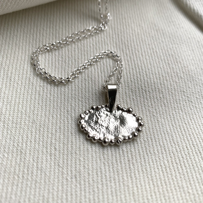 A recycled silver oval pendant with silver granulated details on edges. Displayed on a silver chain on a white cloth backdrop