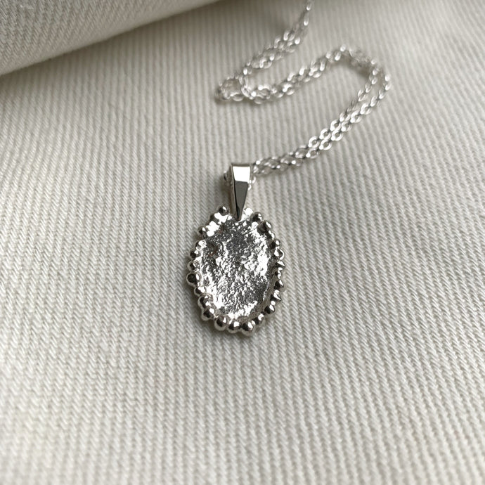 An Oval shaped silver necklace featuring a molten fire textured surface finish with silver beaded edge details.