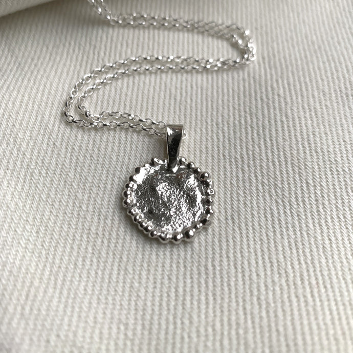 A silver coin style textured pendant necklace embellished with tiny silver balls on the edges. displayed on a white cloth background with a silver chain.