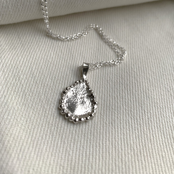 A teardrop shaped silver pendant with a melted surface texture and embellished granulated edge detail. Shown on a white canvas backdrop, hung on a silver chain